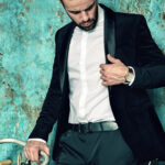 portrait of handsome fashion stylish hipster lumbersexual businessman model dressed in elegant black suit in  posing near sport bicycle and blue grange wall in studio. Metrosexual